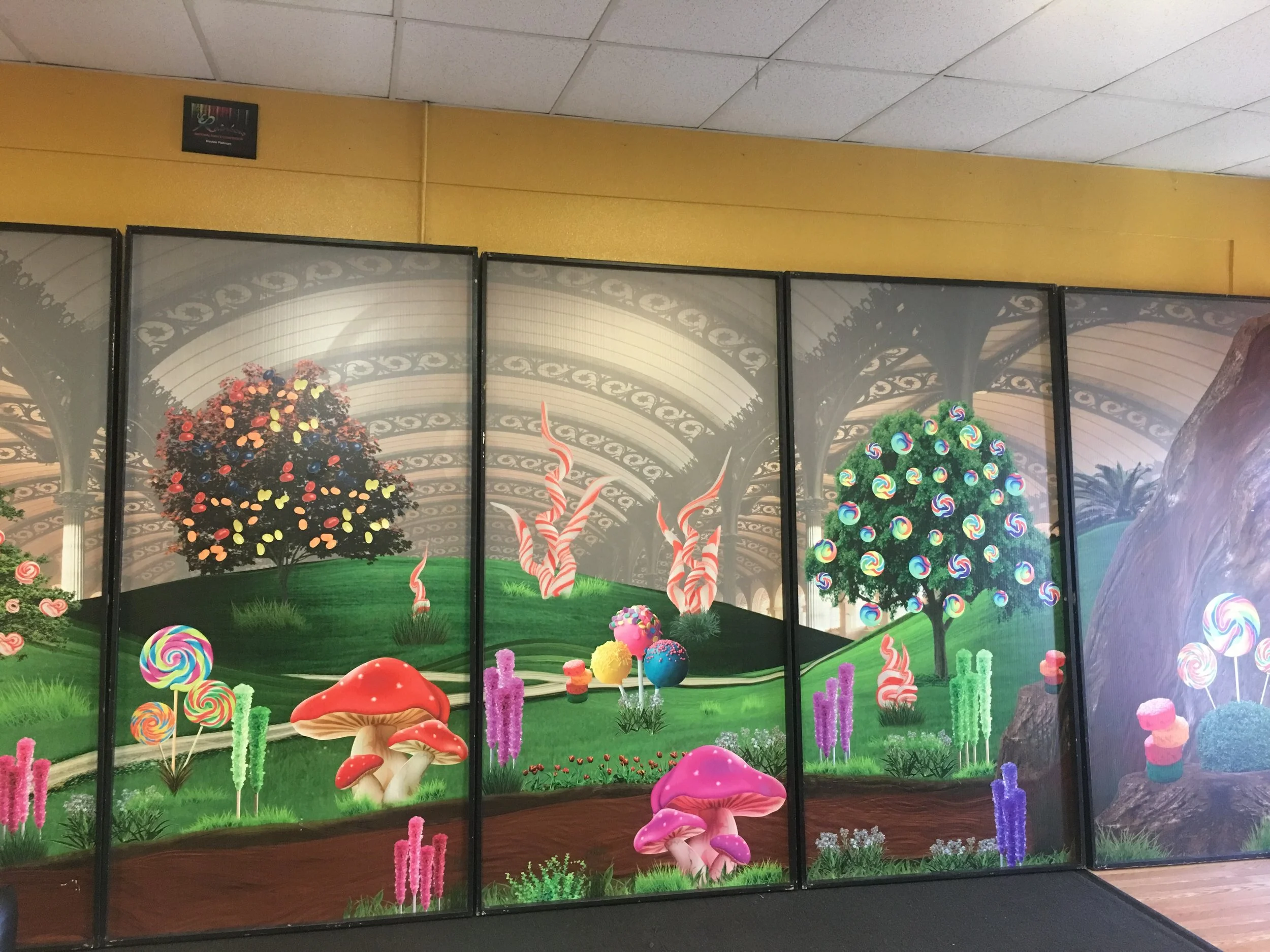 Colorful candy-themed display with decorated trees, mushrooms, and lollipop ornaments, set in front of a fantasy landscape backdrop inside a display case.