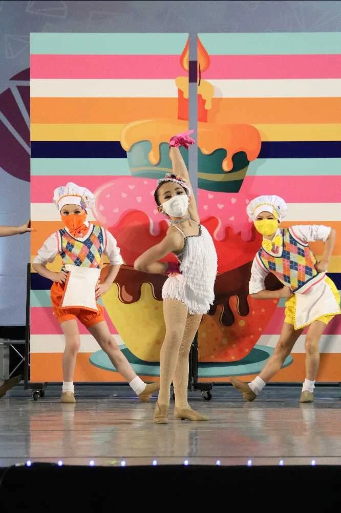 Three young dancers in colorful costumes and face masks perform on stage in front of a large, vibrant backdrop of a cupcake with pink, orange, yellow, and blue frosting and a lit candle.