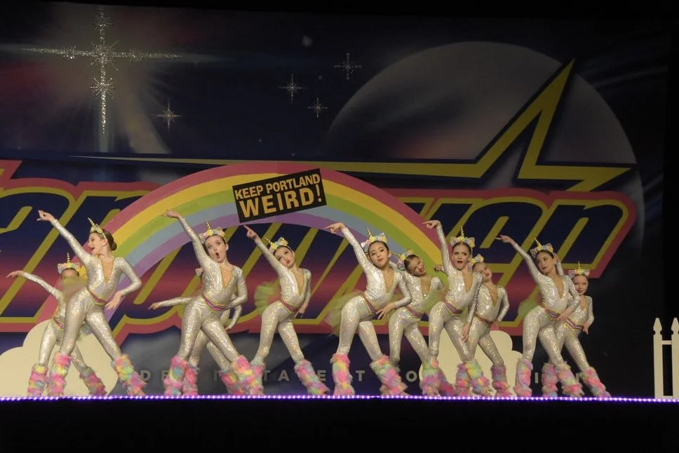 A group of young girls dressed as unicorns performing a dance on stage, with a colorful rainbow backdrop and a sign that reads "Keep Portland Weird!"