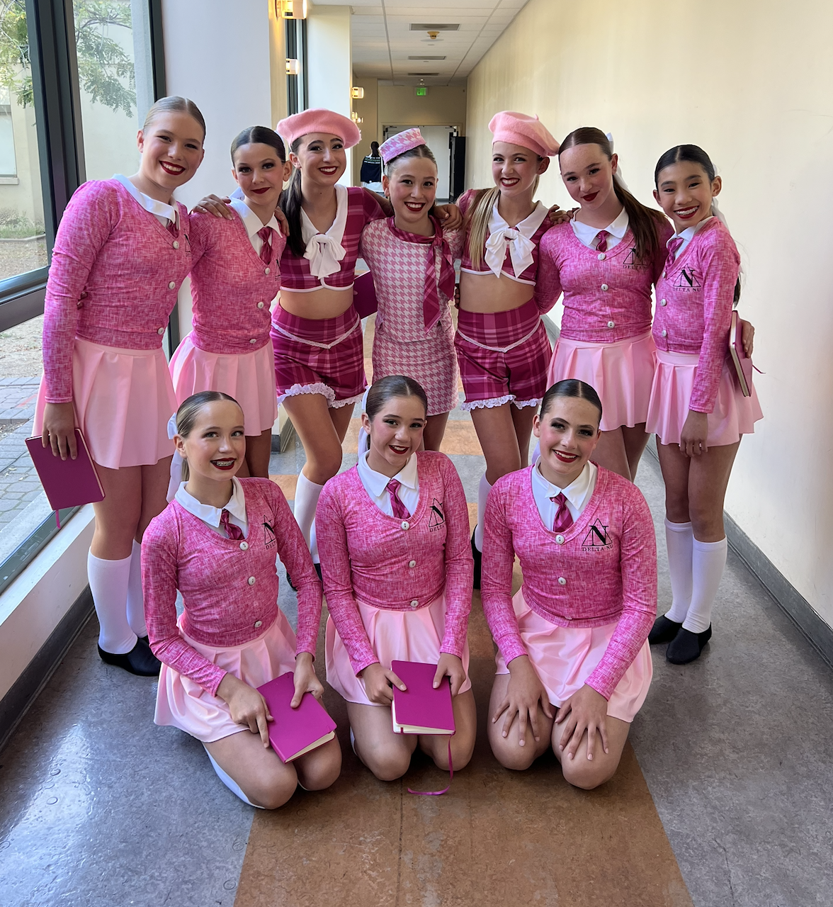 A group of ten young women dressed in pink and white cheerleading or dance costumes, posing together in an indoor hallway.