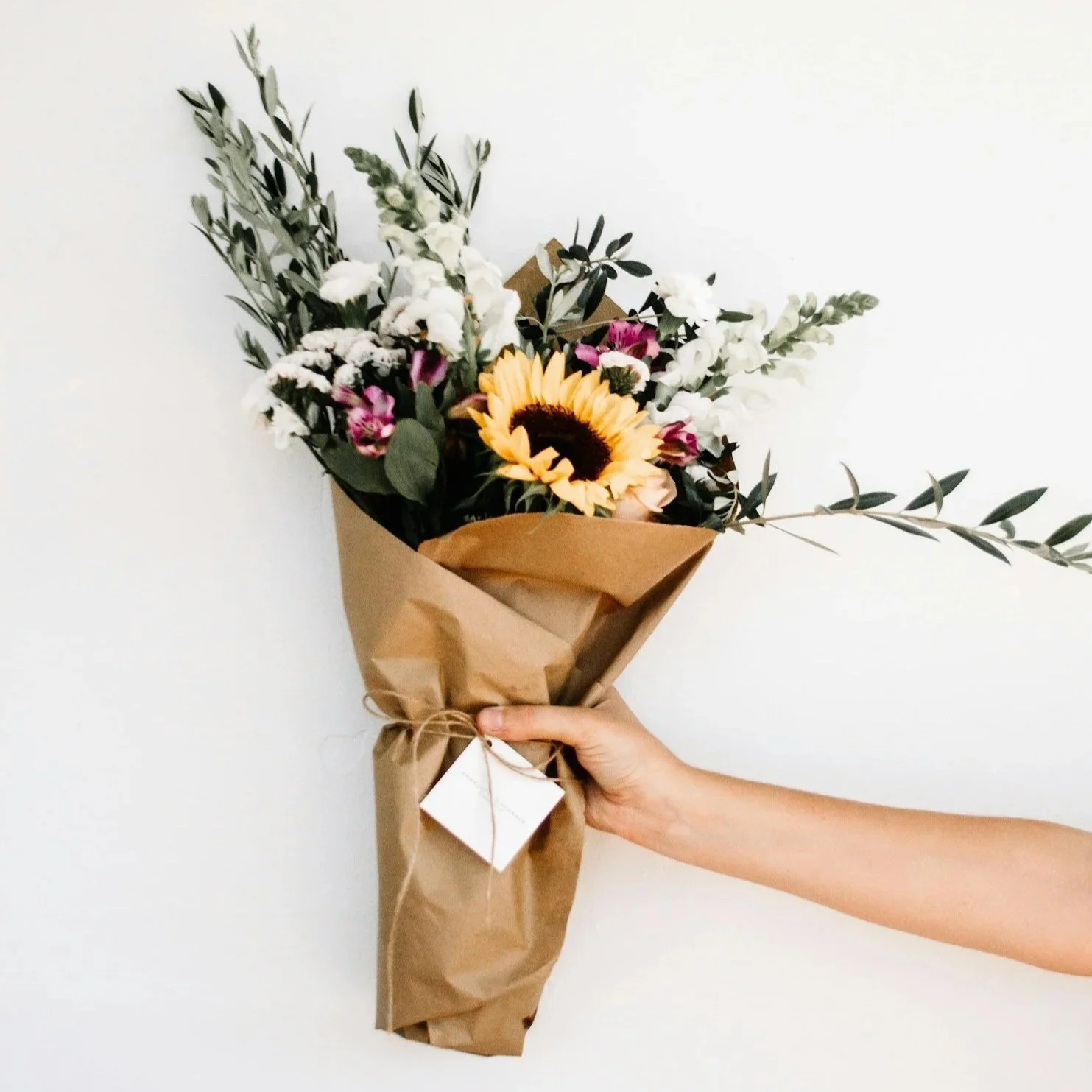 Hand holding a bouquet of flowers wrapped in brown paper, including a sunflower, white and pink blooms, and green foliage.