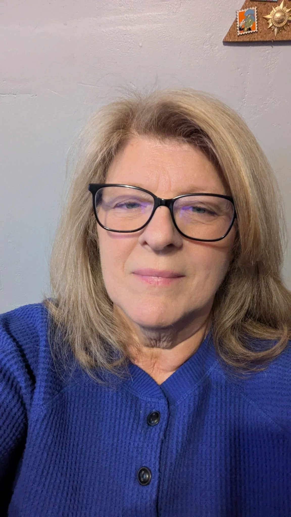 A woman with shoulder-length blonde hair and black glasses wearing a blue sweater, looking at the camera in a close-up selfie.
