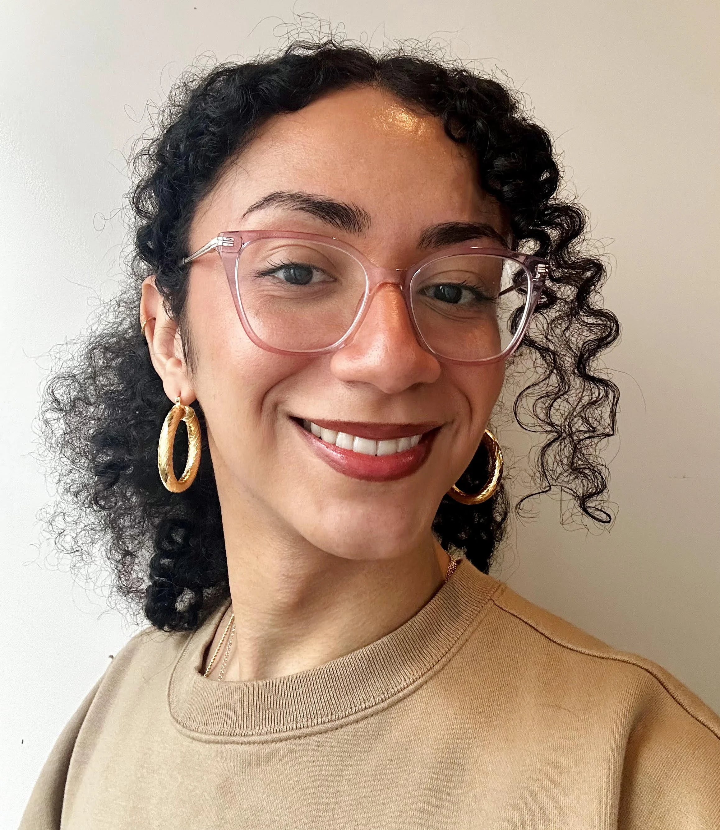 Close-up of a smiling woman with curly dark hair, wearing pink glasses, gold hoop earrings, and a beige sweatshirt, standing against a plain background.