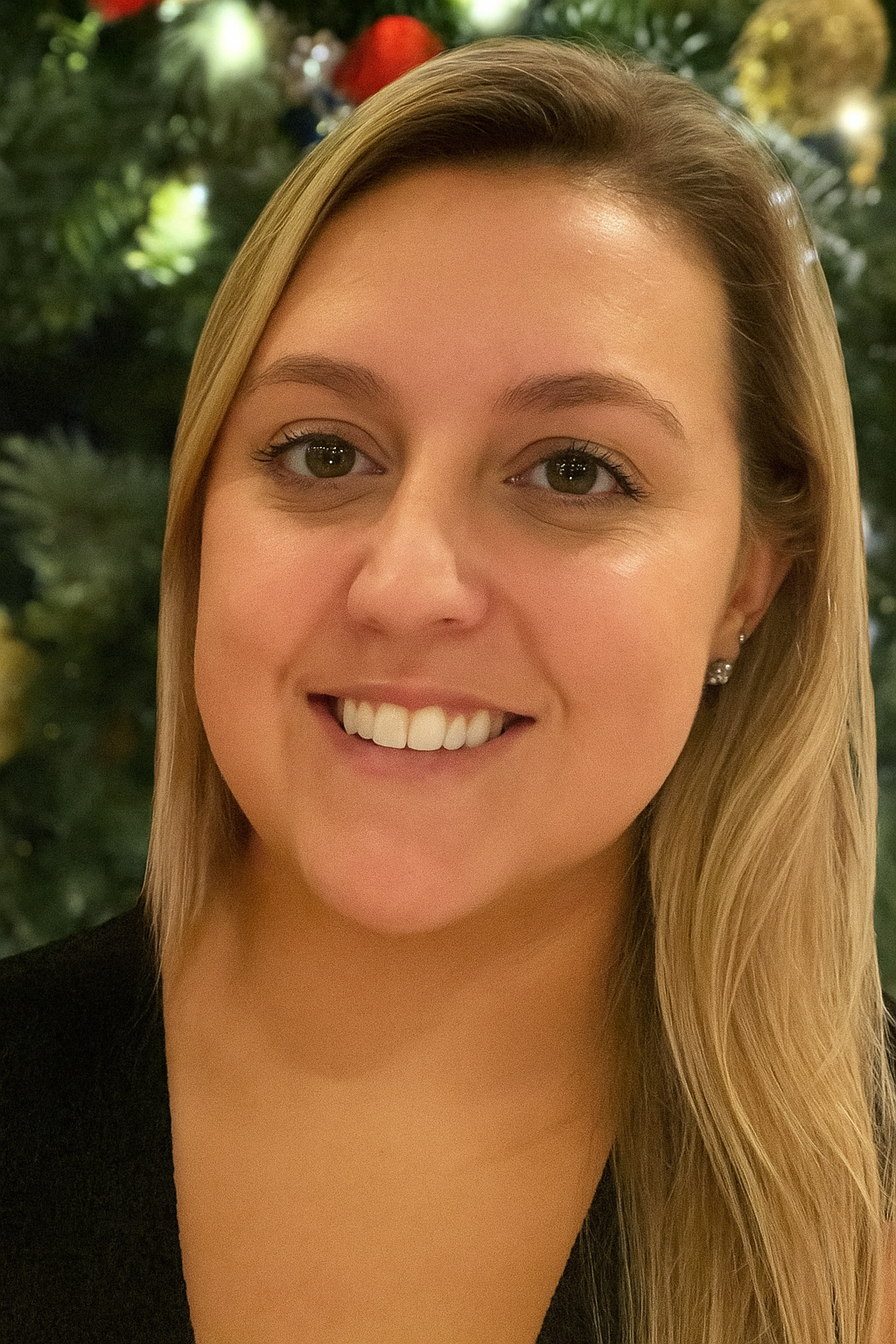 Close-up of a smiling young woman with blonde hair and light makeup, wearing earrings, and a black top, with green foliage in the background.