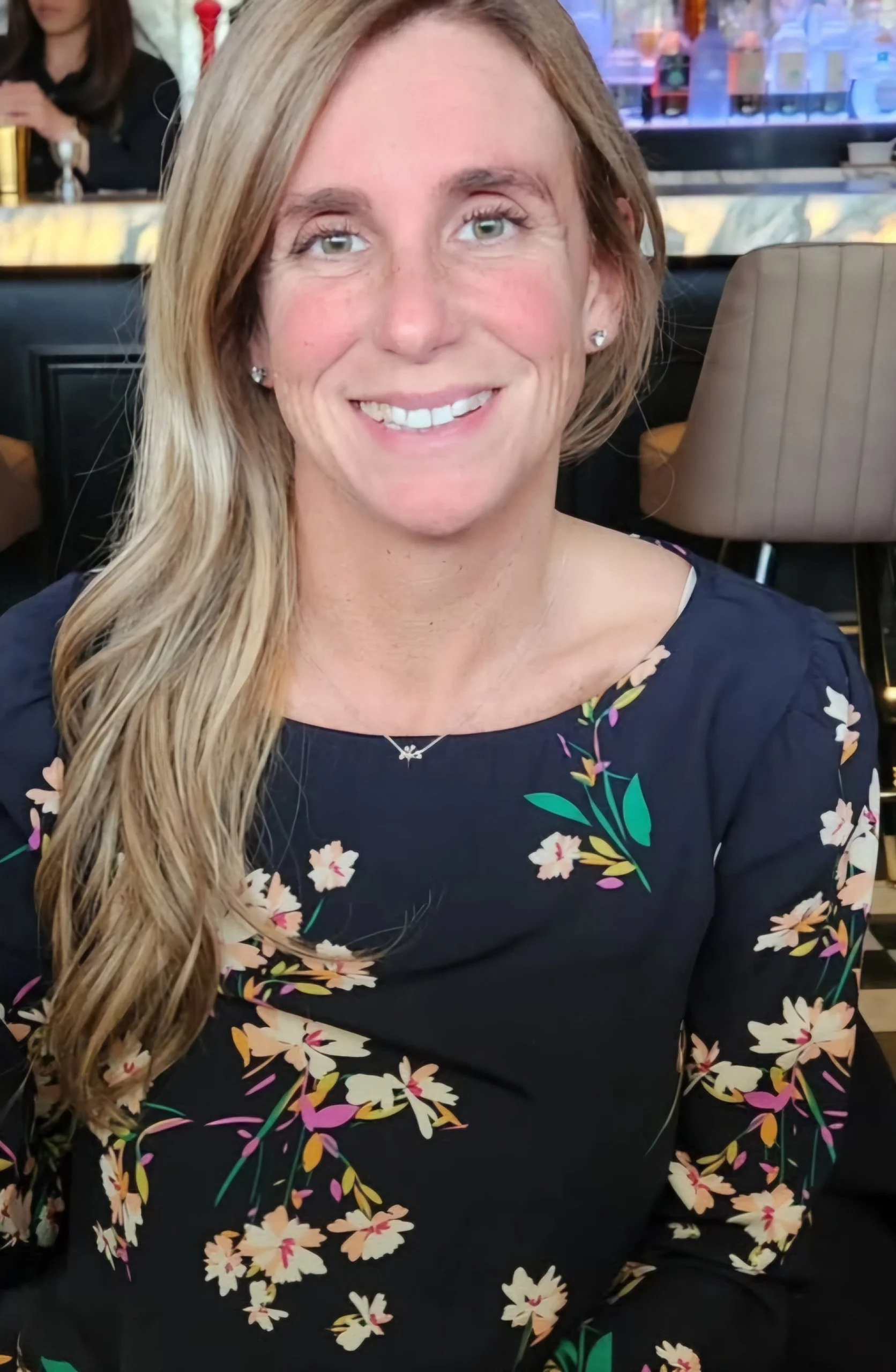 Close-up of a woman with long blonde hair, wearing a black dress with a floral pattern, smiling at the camera, in a restaurant or cafe setting.