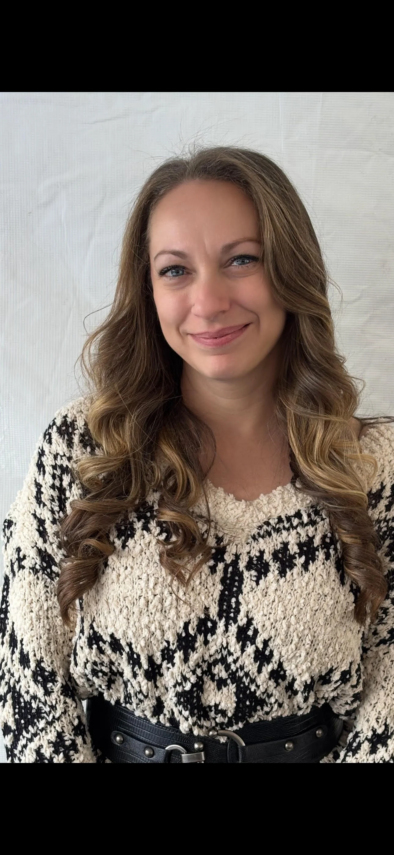A woman with long wavy brown hair and blue eyes wearing a black and white patterned sweater and a black belt, standing in front of a plain white background.