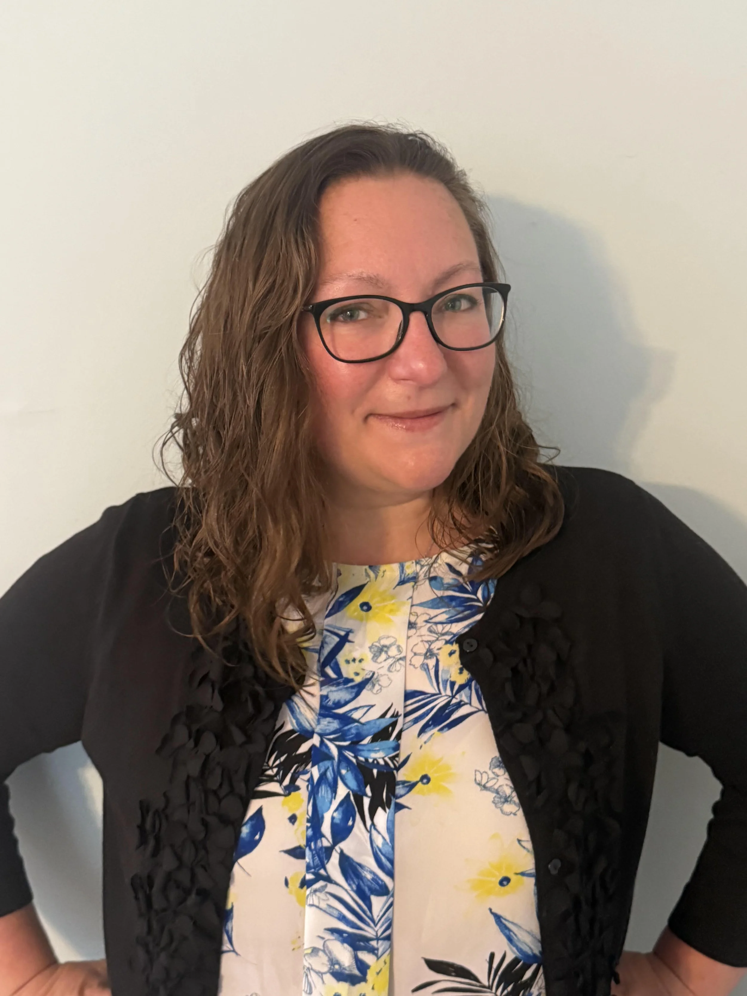 Woman with wavy brown hair, glasses, wearing a black cardigan over a floral dress with blue, yellow, and white colors.
