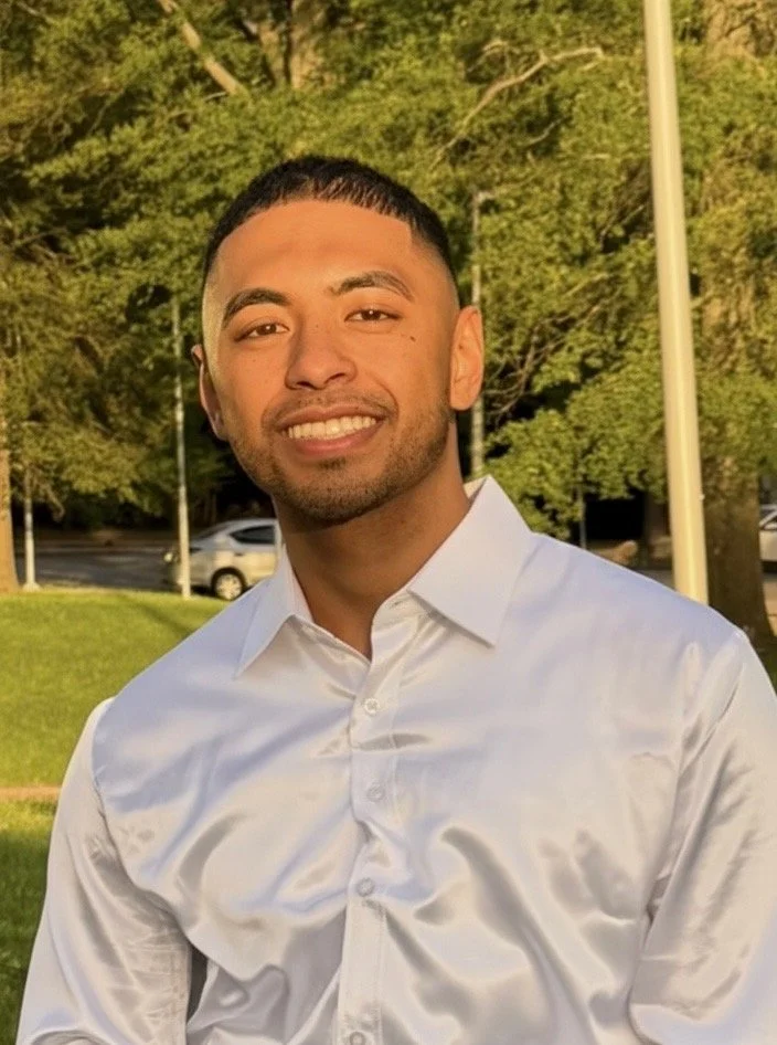 A young man with short black hair, smiling, wearing a white dress shirt outdoors with trees, cars, and a lamppost in the background.