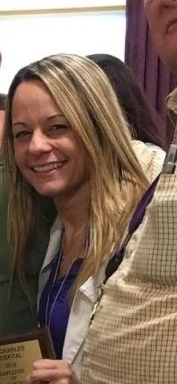 Smiling woman with blonde hair and doctor's coat, holding a plaque, standing next to a person in a checked shirt.