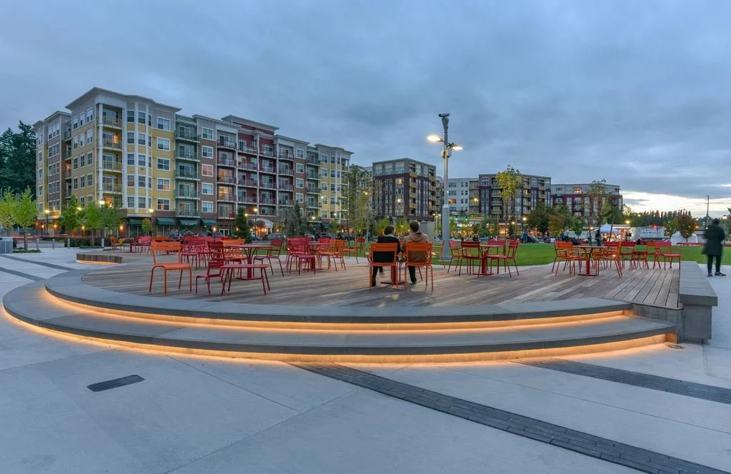 An outdoor public space at dusk with a wooden stage, red and orange chairs, and apartment buildings in the background.