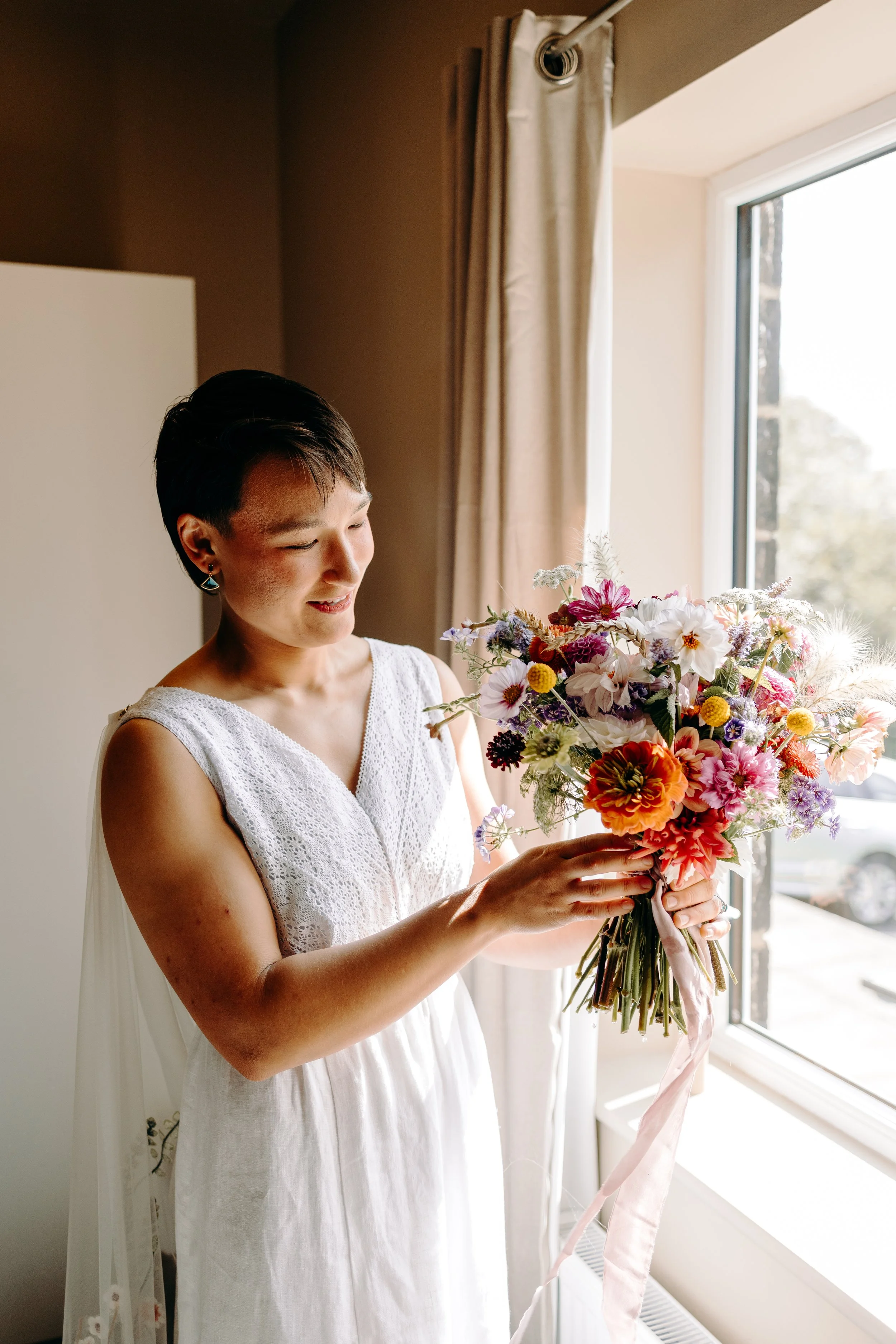 A woman in a white dress holds a colorful bouquet of flowers near a window, illuminated by natural light.