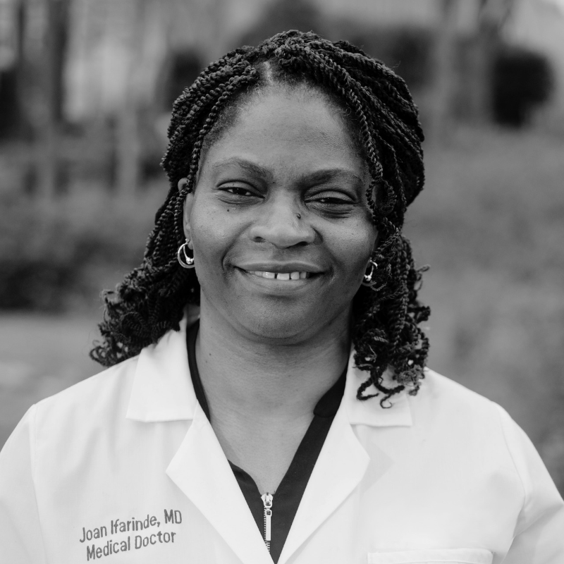 Black and white photo of a smiling female medical doctor with twisted hair, wearing a white coat with her name Joan Ifarinde and 'MD Medical Doctor' embroidered on it.