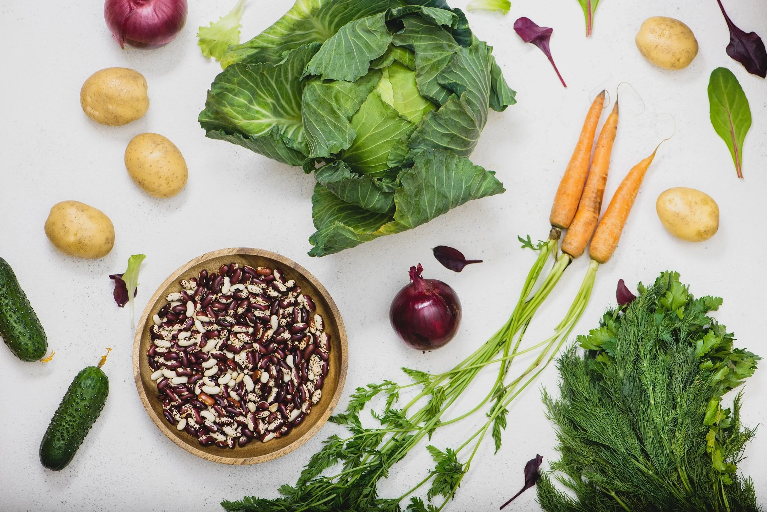 Assorted fresh vegetables including a head of green cabbage, three carrots with green tops, a pile of small potatoes, two cucumbers, a red onion, leafy greens, and a bowl of mixed beans, all on a white surface.