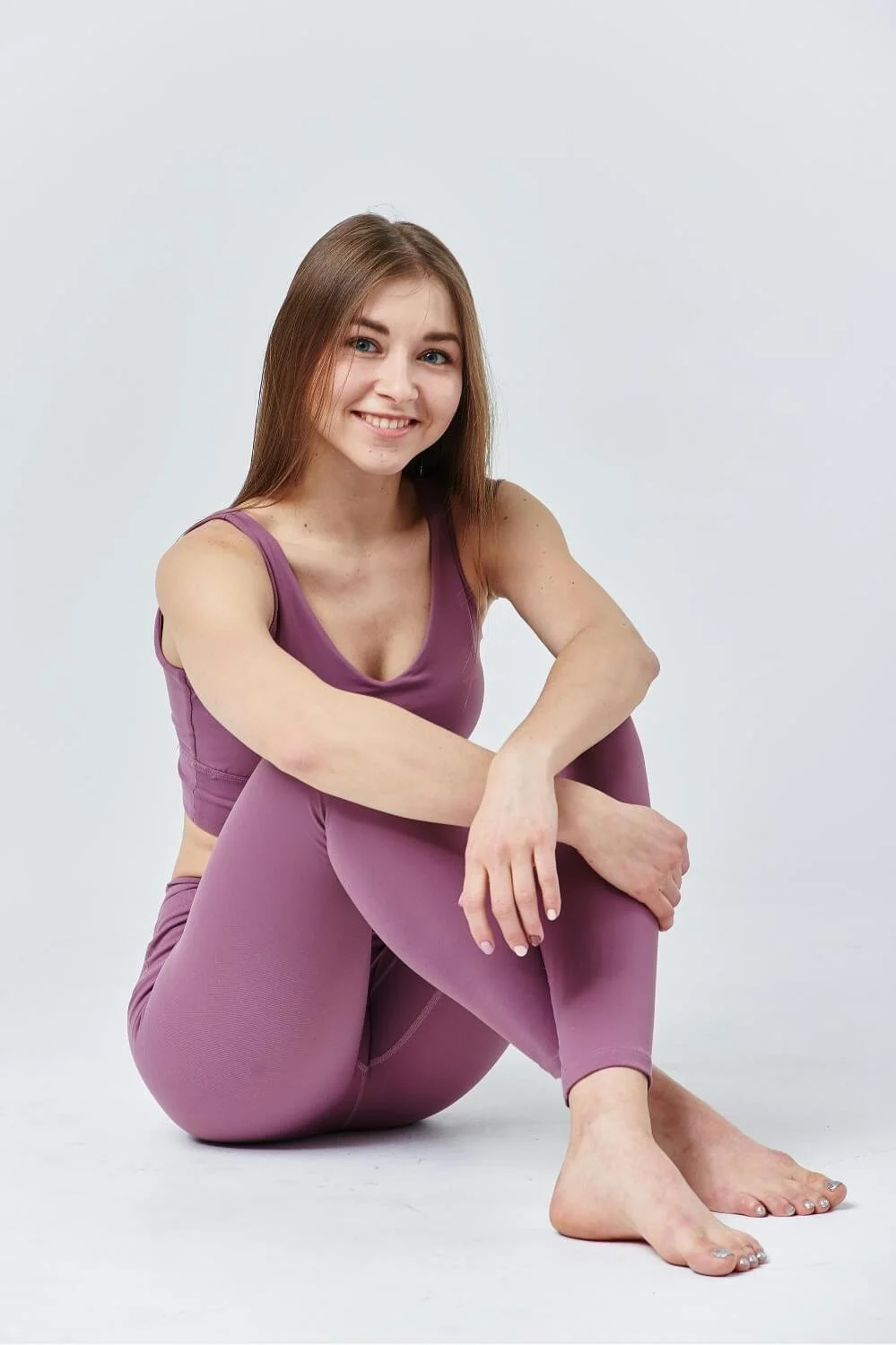 A young woman with long brown hair sitting on the floor against a plain light background. She is smiling and dressed in matching purple workout attire, with her legs crossed and arms resting on her knees.