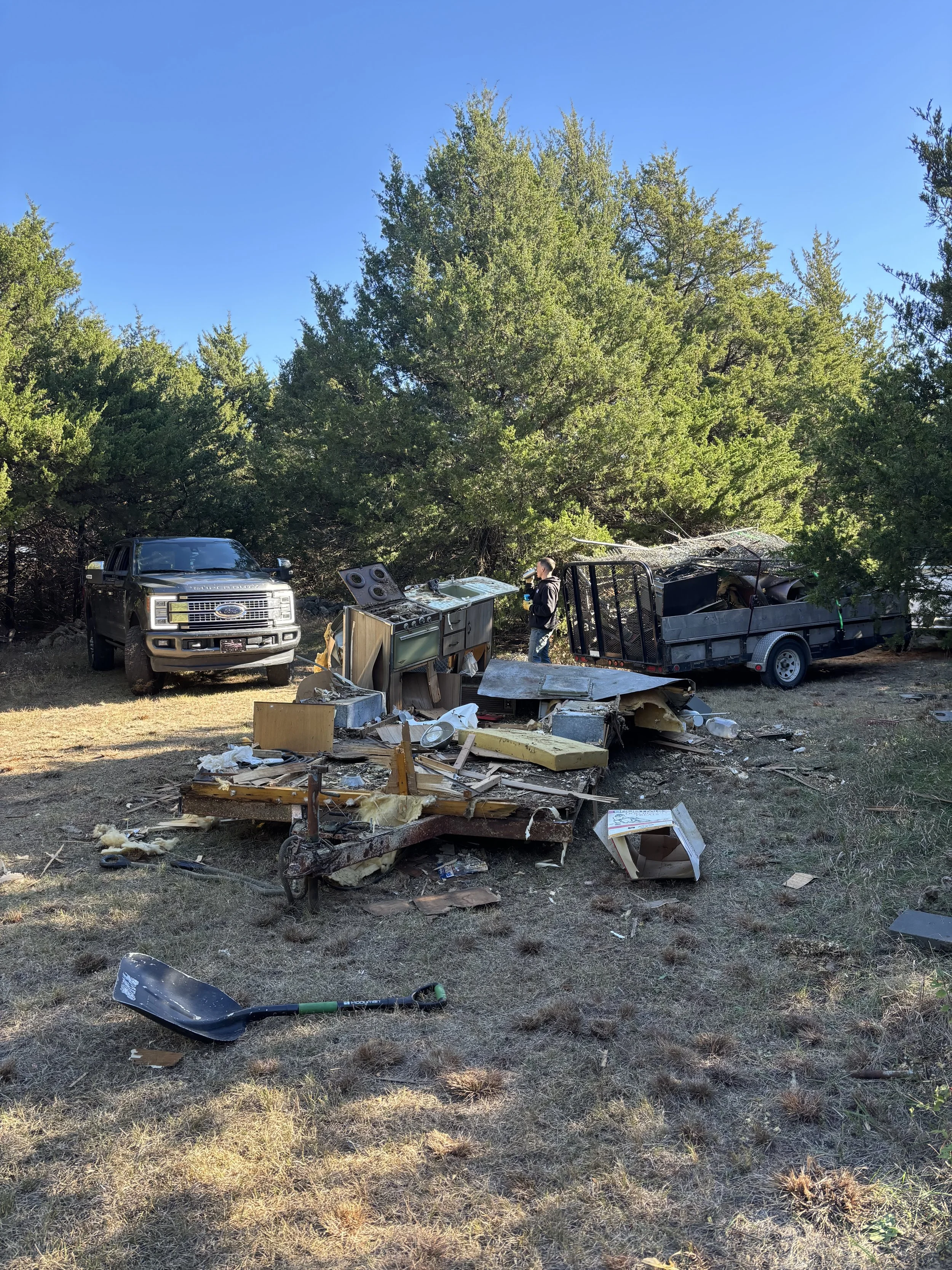 Pile of discarded furniture including white cabinets, wooden crates, and chairs stacked outside on grass.