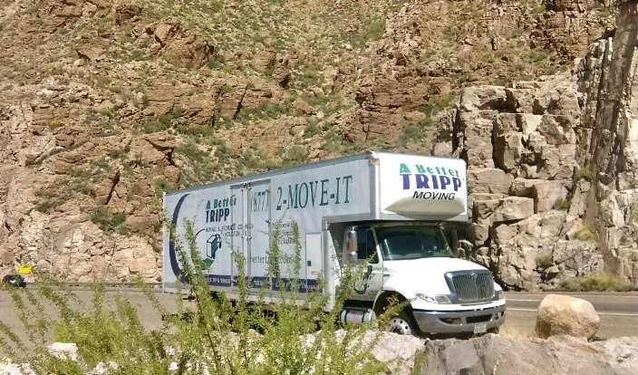 A white delivery truck driving on a curved two-lane road surrounded by green trees and hills, under a blue sky with some white clouds.