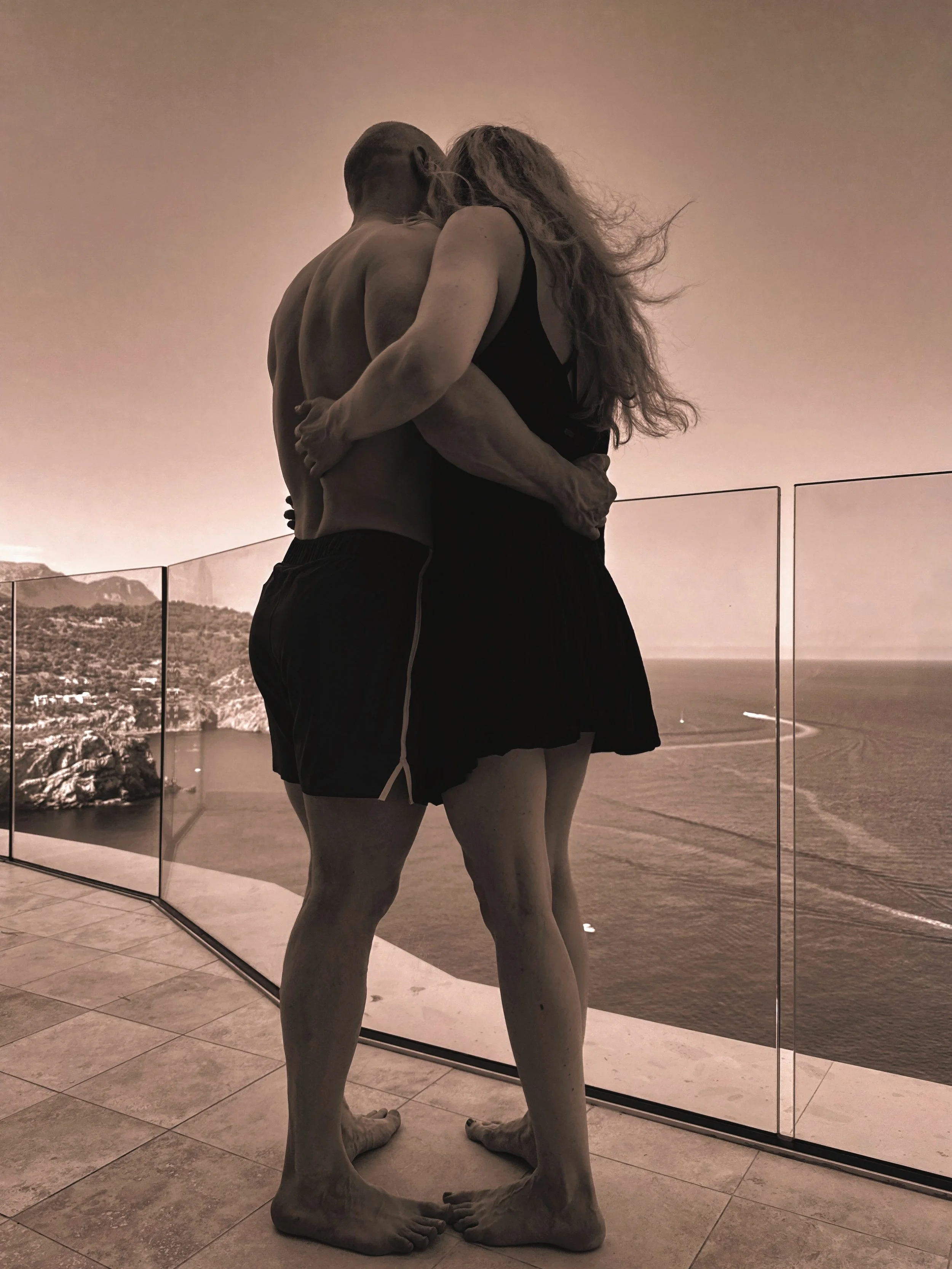 Statue of two people embracing, standing with their faces close together, in front of a large window with a scenic ocean view.