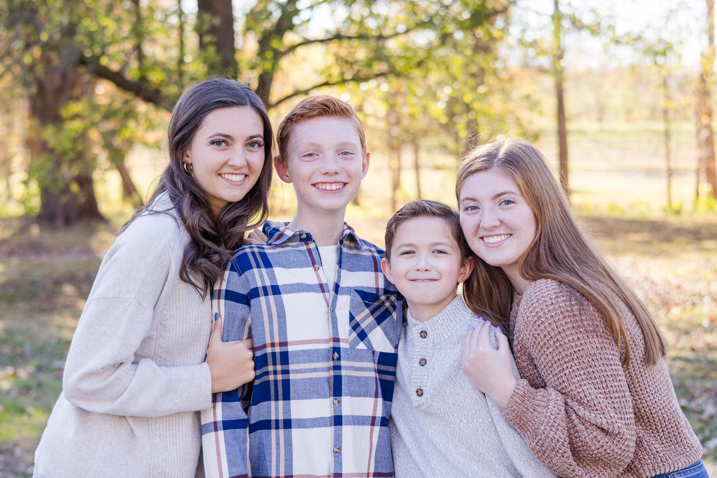Four children smiling outdoors among trees with autumn foliage.