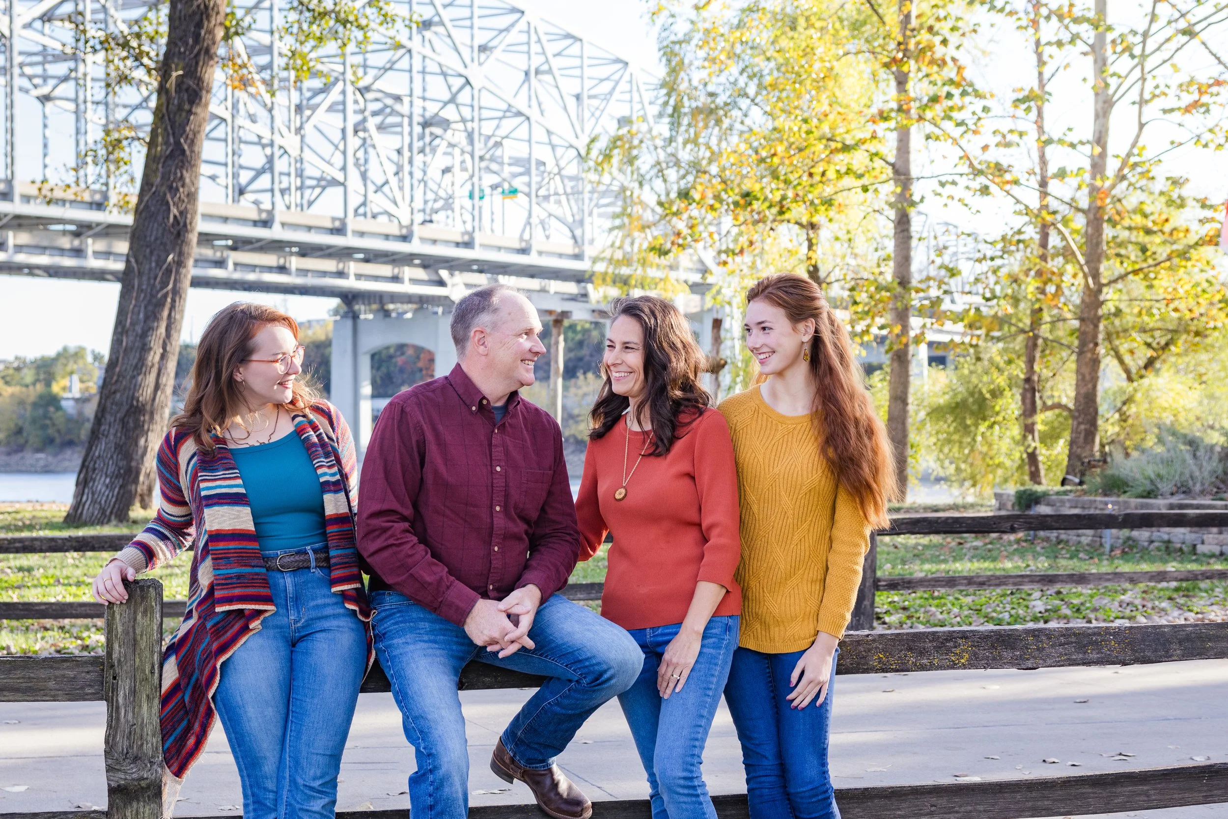A group of four people, two women and two men, standing and sitting on a wooden fence in a park with autumn foliage and a bridge in the background, smiling and talking.