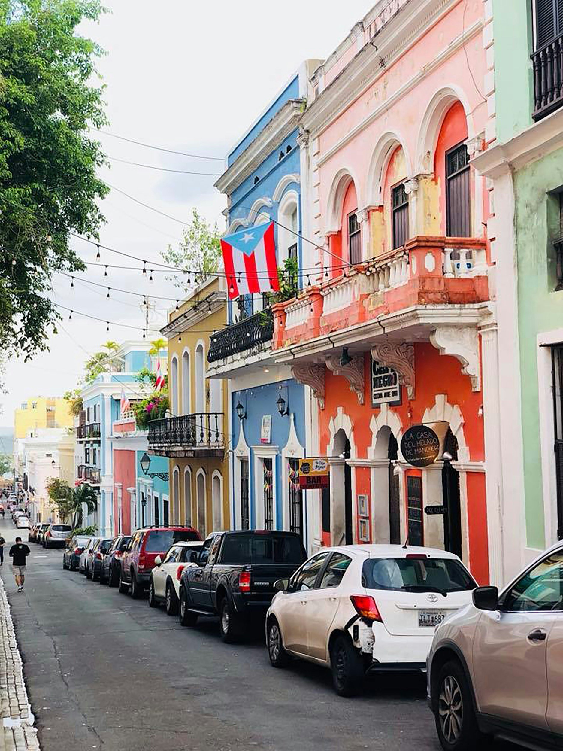 Colorful buildings lining a street with parked cars and string lights, with a Puerto Rican flag hanging above.