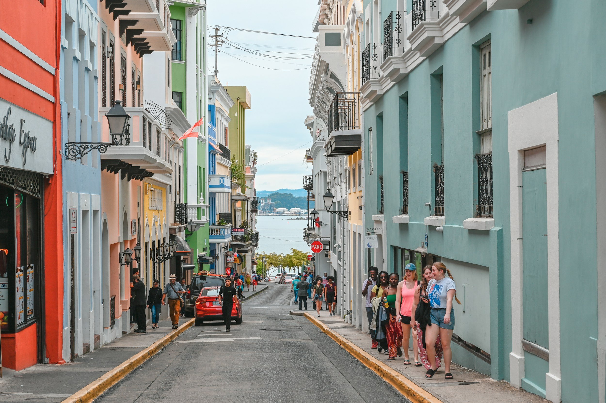 Colorful buildings line a busy street with pedestrians walking on the sidewalk and a view of a body of water in the background.