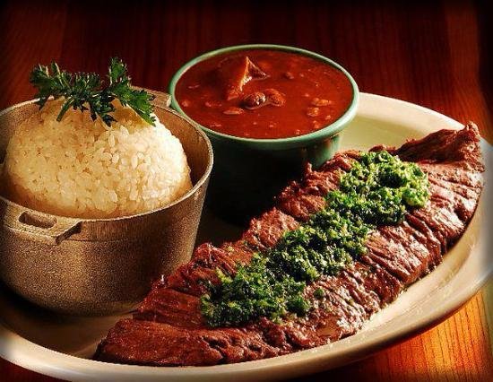 Plate of grilled steak topped with chimichurri, white rice garnished with parsley, and a bowl of red sauce on a wooden table.