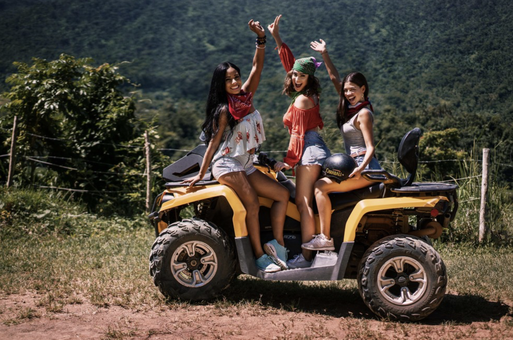 Three smiling women on a yellow all-terrain vehicle in a rural outdoor area with green hills in the background.