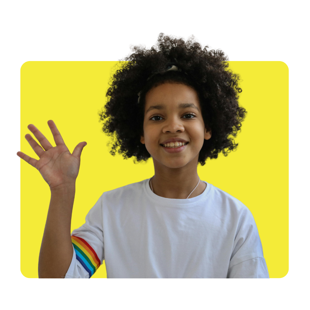 Smiling young girl with curly hair wearing a white t-shirt with rainbow stripes on the sleeve, waving and standing in front of a yellow background.
