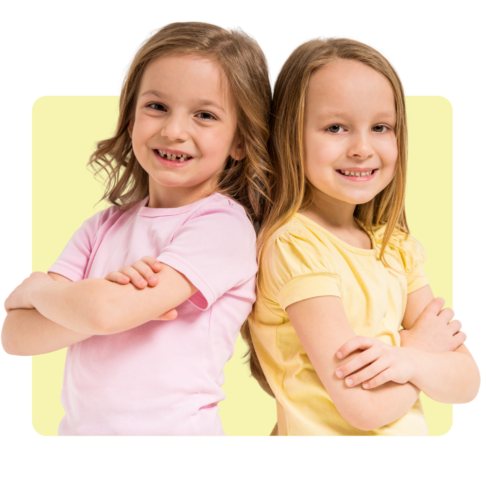 Two young girls with light brown hair, smiling and standing with crossed arms against a light yellow background.