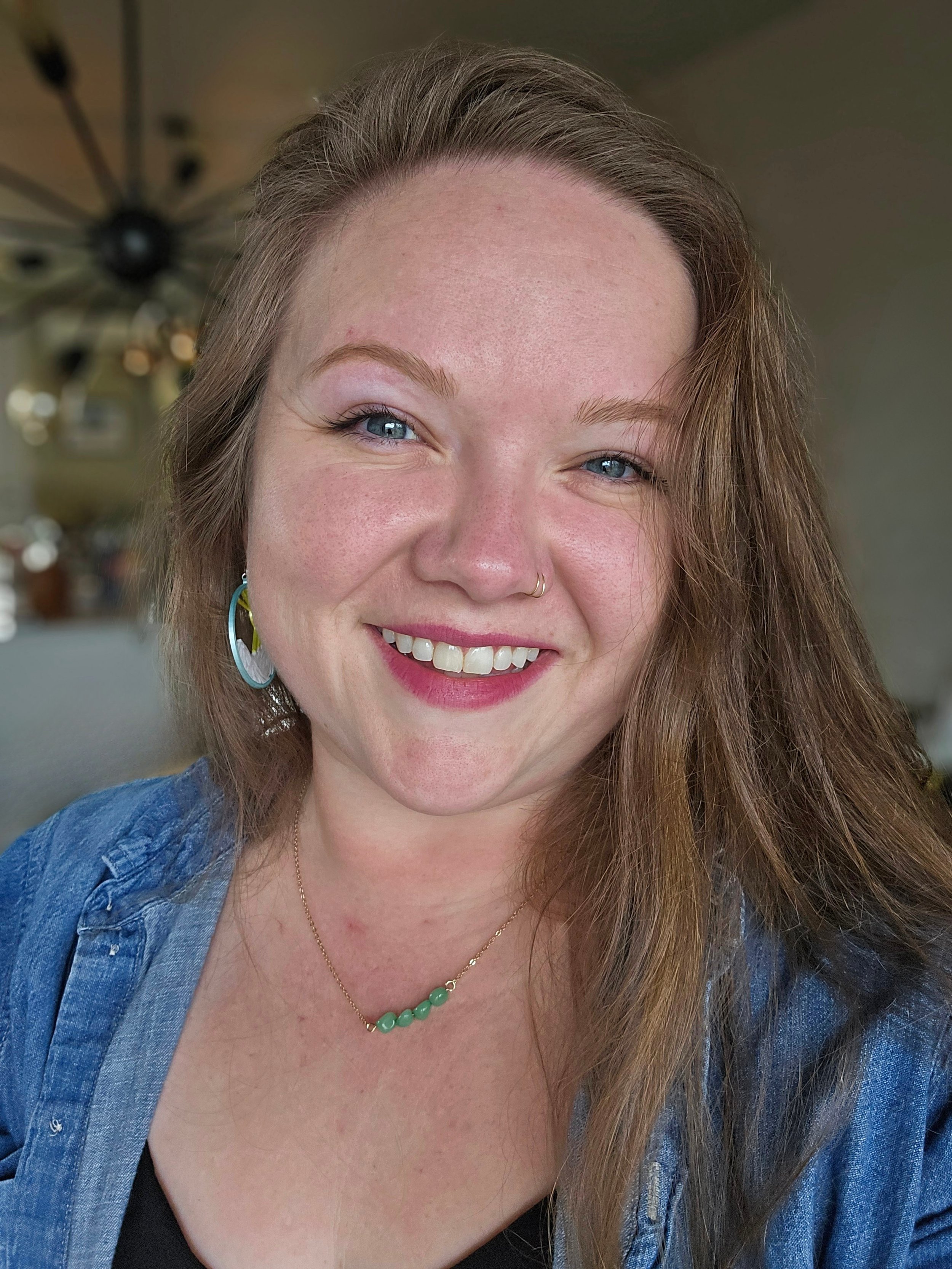 Close-up of a smiling woman with blue eyes, freckles, long brown hair, wearing a denim jacket, a gold chain with green beads, and silver earrings, in a well-lit indoor setting.