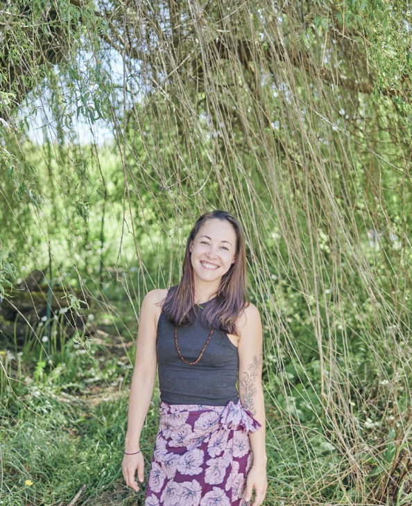 Young woman smiling outdoors, standing under a large tree with hanging branches, surrounded by green foliage.