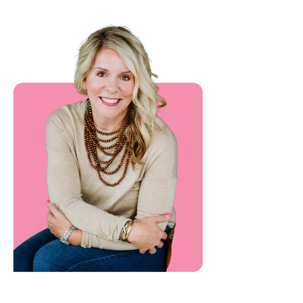 Smiling blonde woman with curly hair, wearing a beige sweater, multiple layered necklaces, a watch, gold bracelets, and blue jeans, sitting with arms crossed against a pink background.
