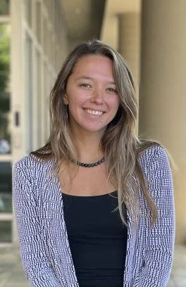 A young woman with long blonde hair smiling, standing outdoors with a building and columns in the background.