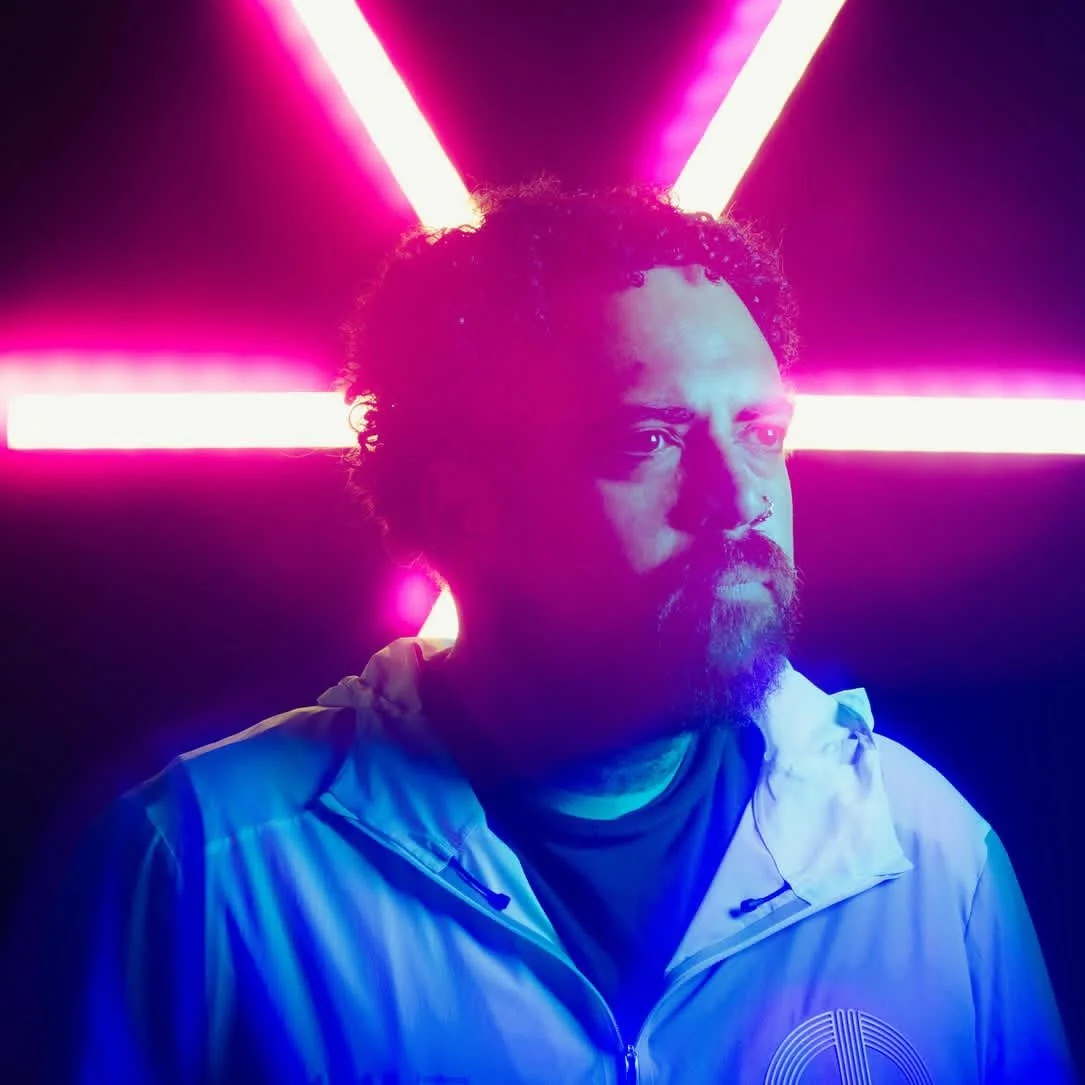 A man with a beard and curly hair standing against a neon pink and purple background.