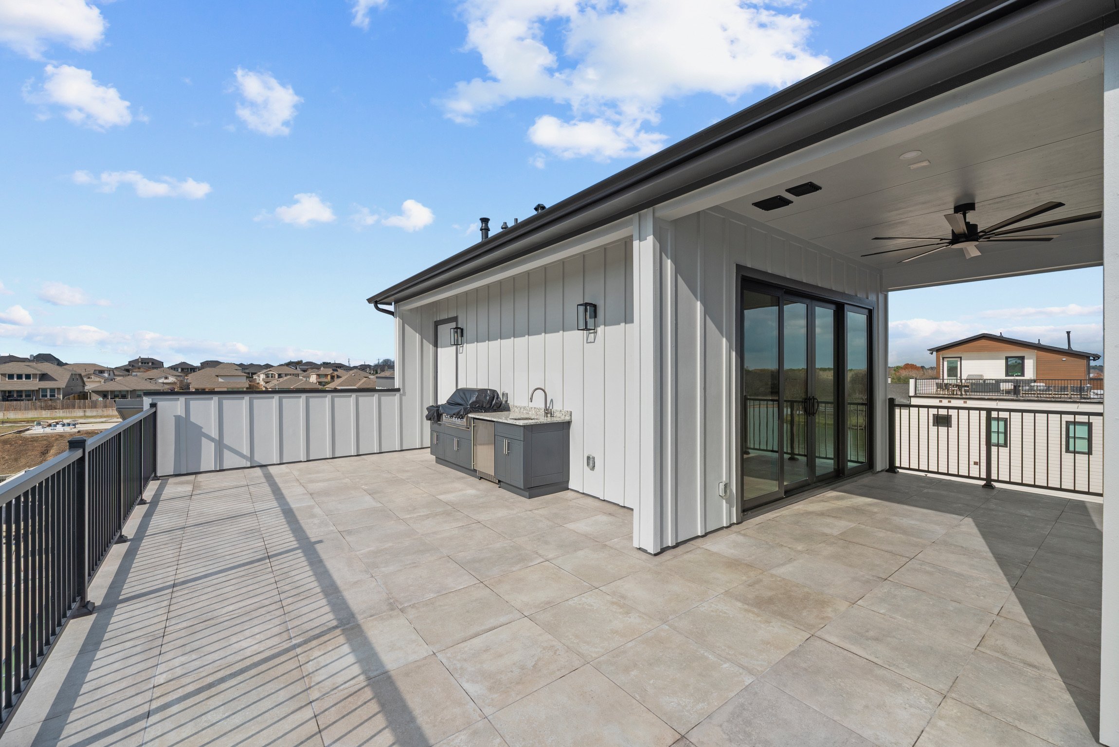 Large rooftop terrace with beige tiled flooring, metal black railing, outdoor kitchen with grill, sink, and storage, and sliding glass door leading inside. Blue sky with scattered clouds overhead.