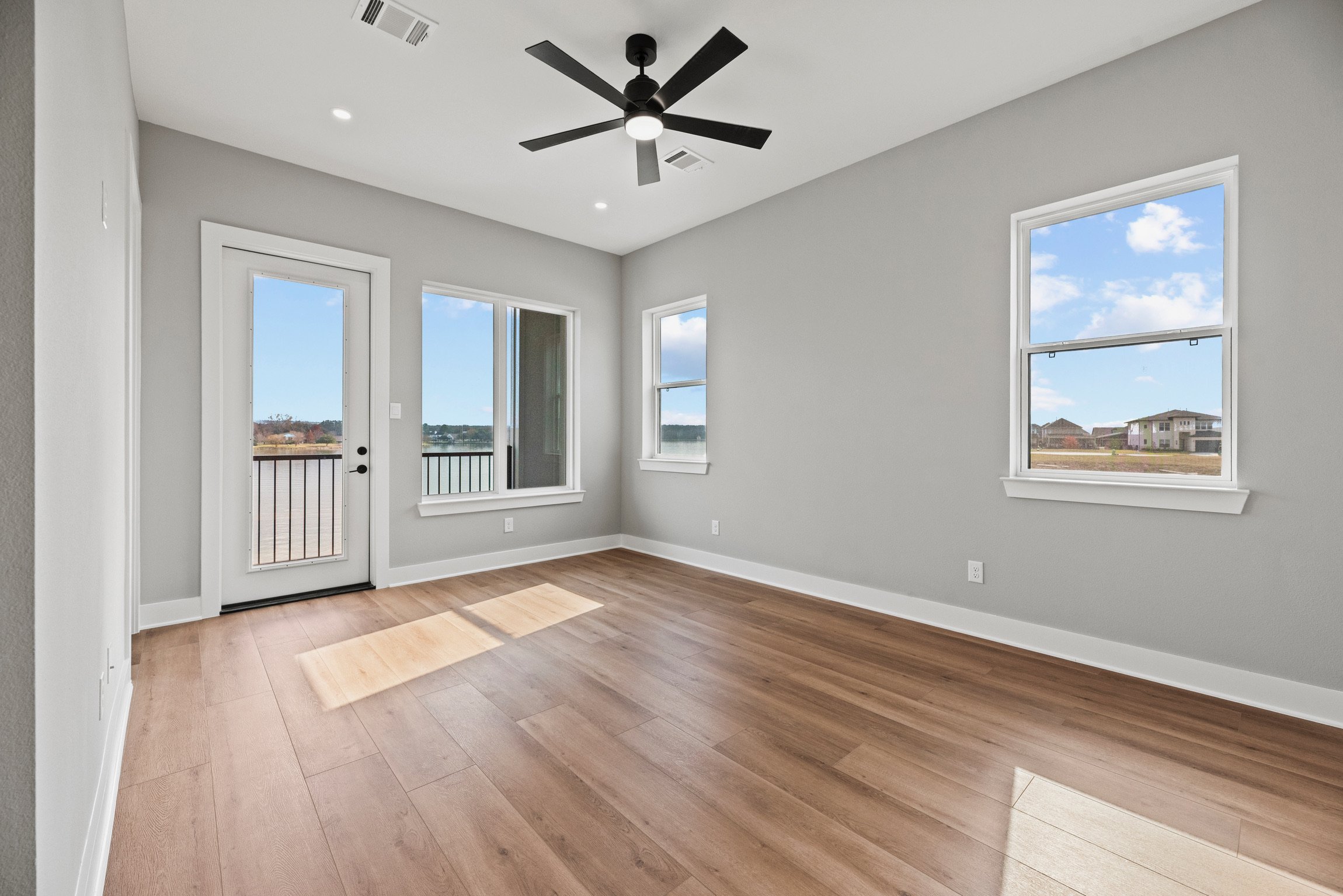 Empty room with hardwood floors, three large windows, a glass door with a screen, white walls, and a black ceiling fan.