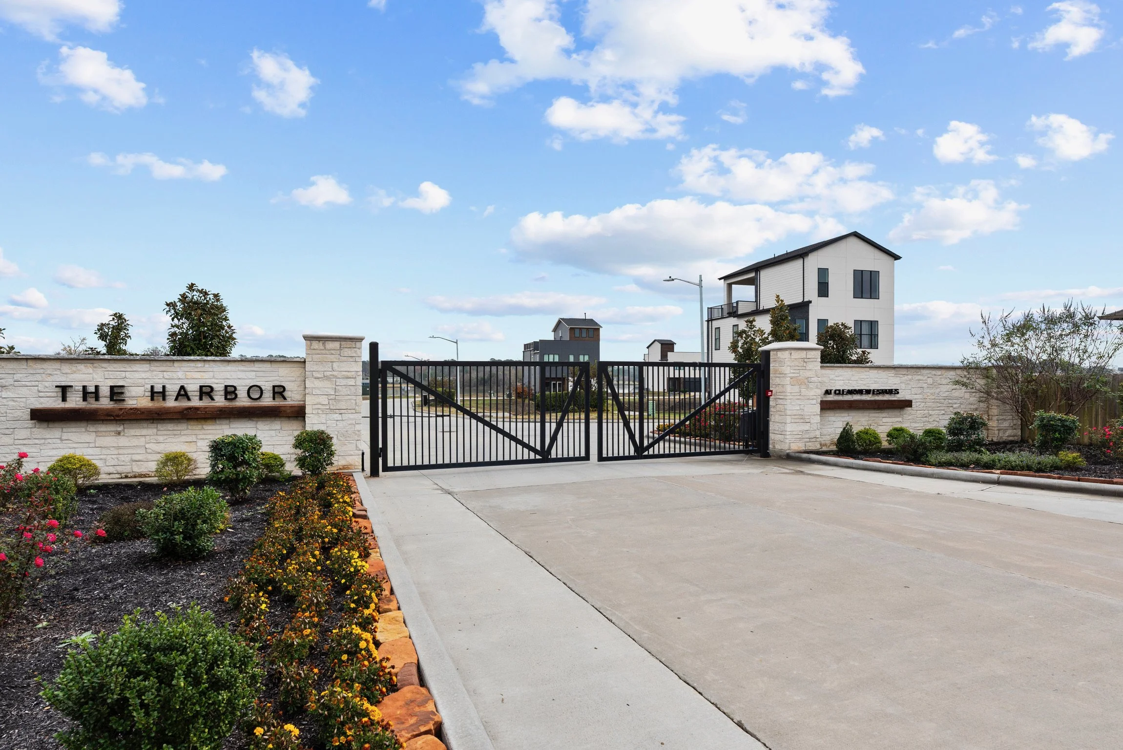 Entrance gate to The Harbor residential community with stone walls, black metal gates, and landscaped flower beds with bushes and small trees under a partly cloudy sky.