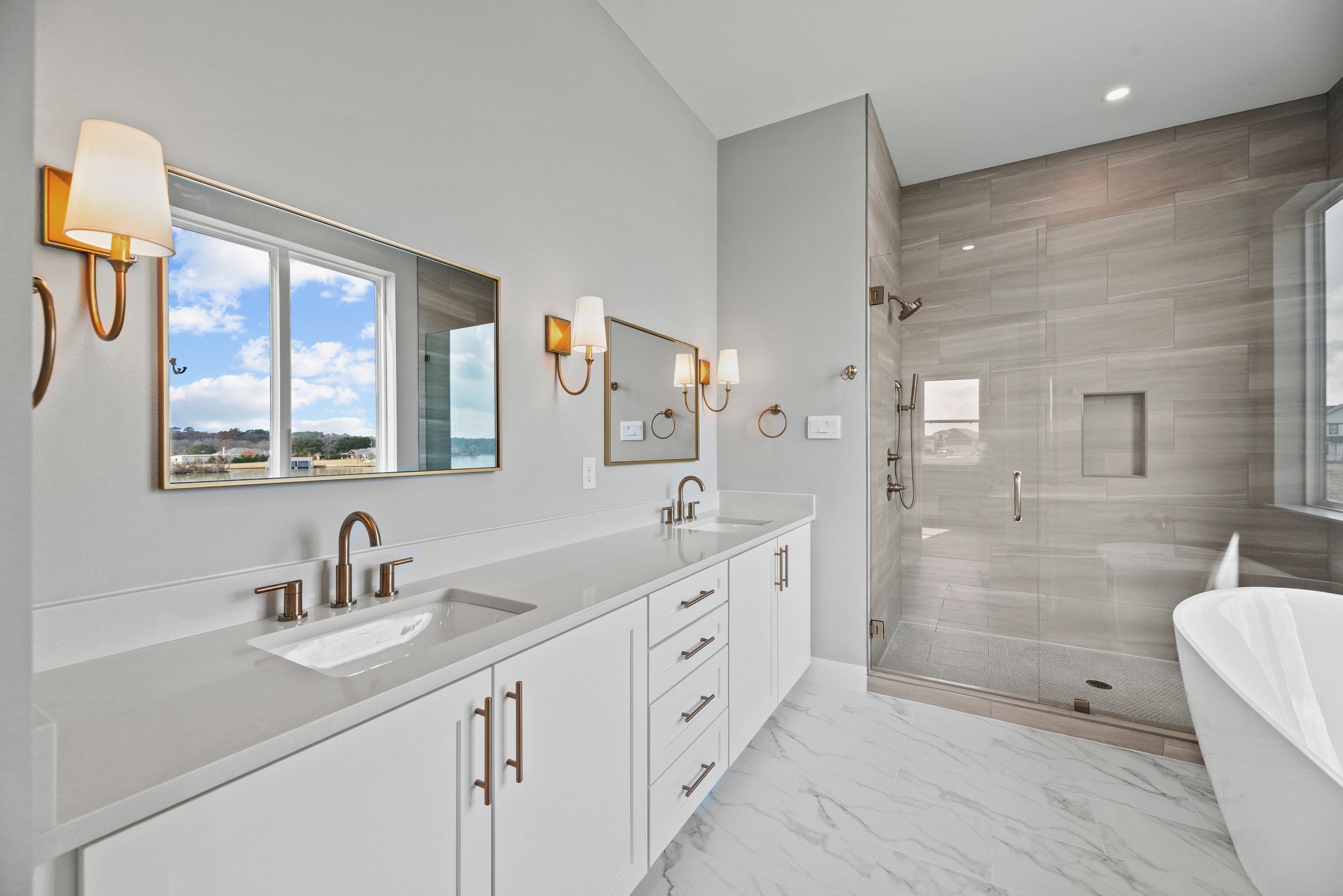 Modern bathroom with double vanity, large mirror, wall-mounted lights, walk-in shower with glass door, bathtub, and window with a view of blue sky and clouds.