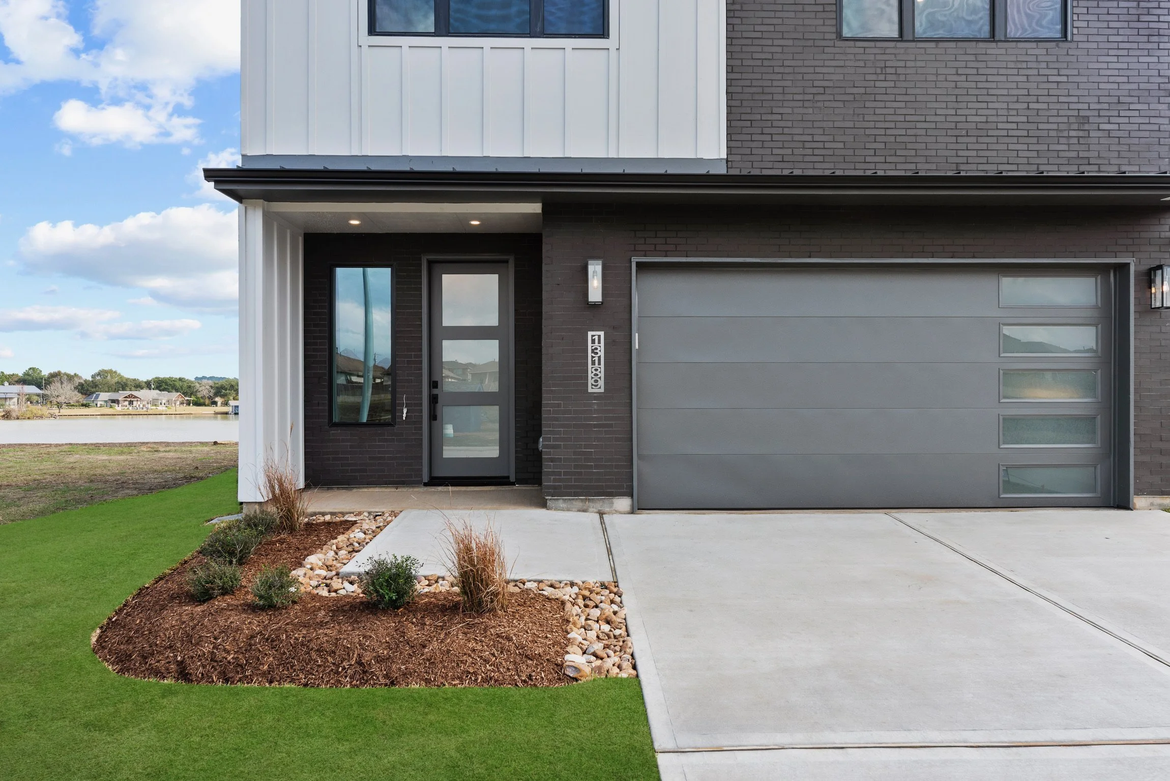 Modern house with a gray front door, a large gray garage door with glass inserts, and a well-maintained lawn with landscaped flower beds.