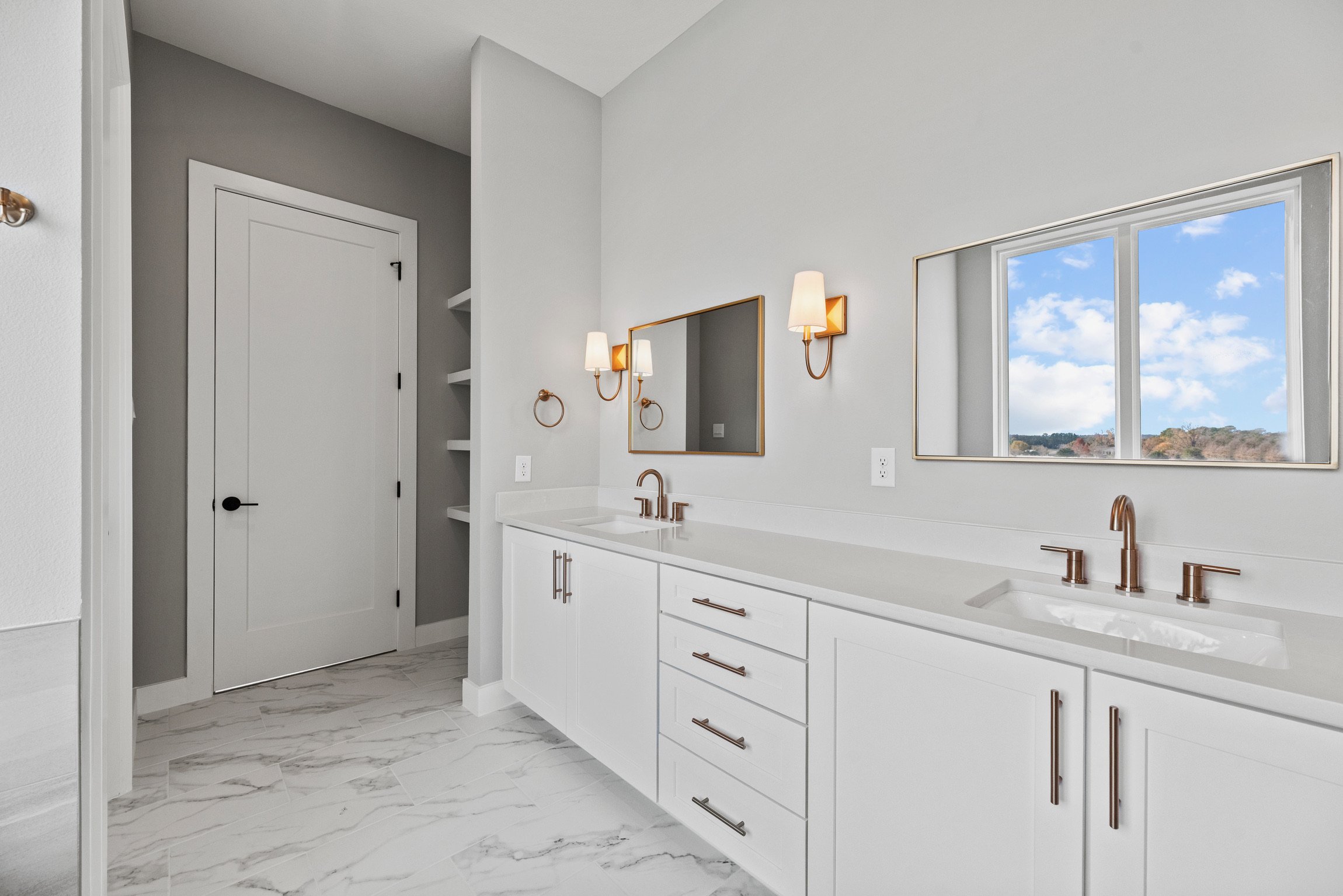 Modern bathroom with dual sinks, white cabinets, gold fixtures, large mirror, window with view, marble floor, and wall sconces.