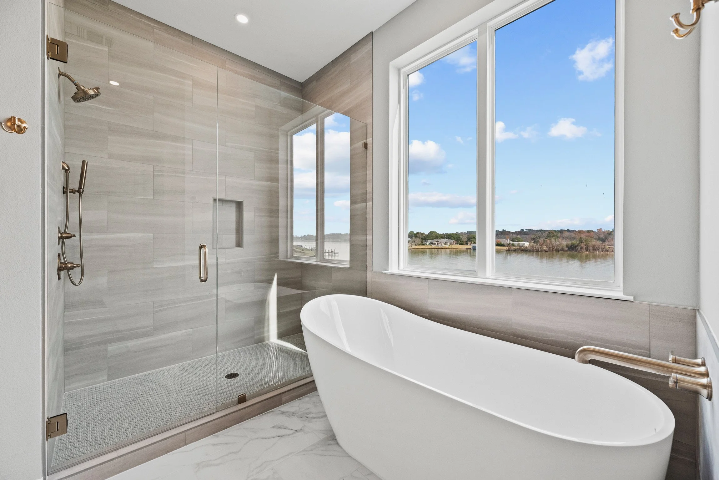 Modern bathroom with a large window overlooking a lake, featuring a walk-in shower with glass doors and a freestanding bathtub.