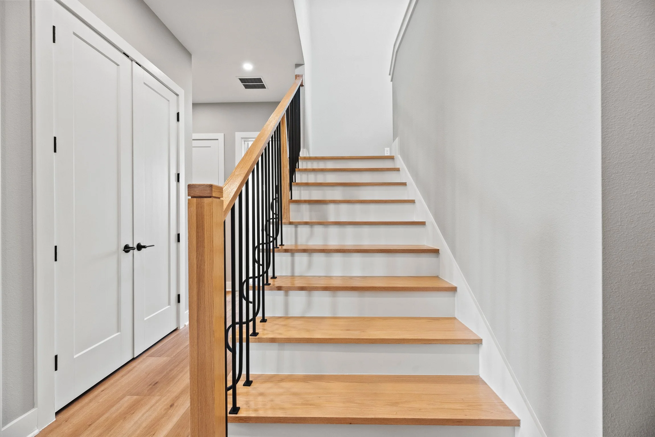 Interior of a modern house with a staircase featuring wooden steps, black metal balustrades, and white walls, adjacent to a closet with two white doors.