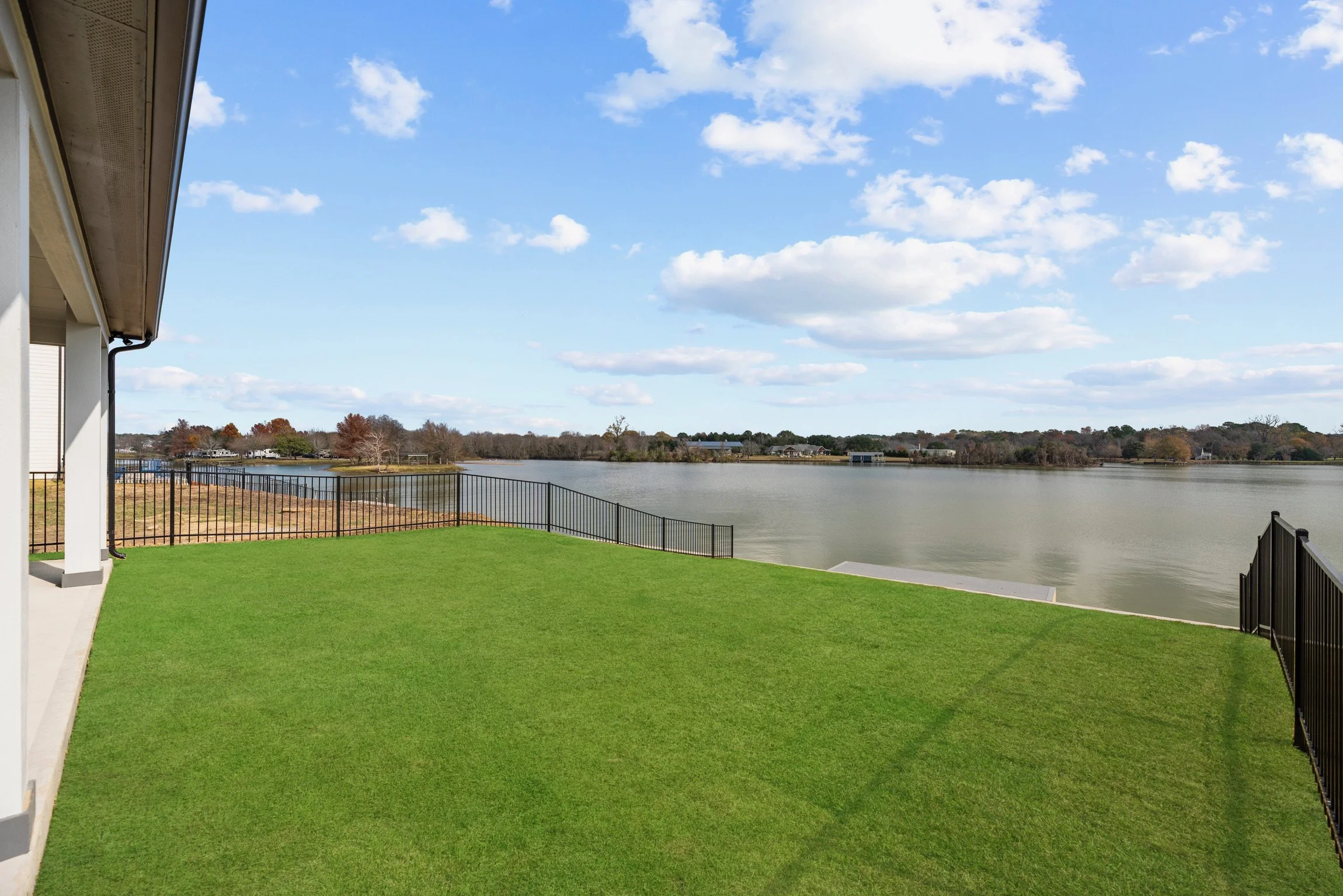 View of a backyard with green grass, black metal fencing, and a lake under a blue sky with clouds.