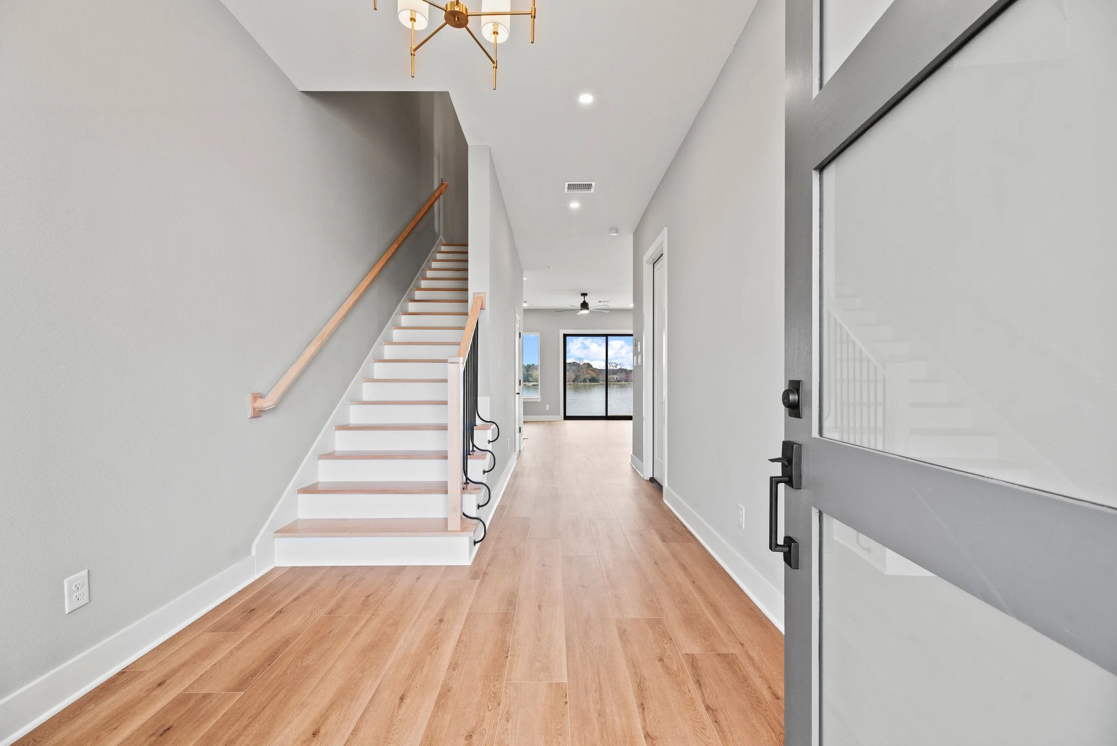 Empty modern house entryway with light wood flooring, a staircase with a wooden handrail, and a view of a living area with sliding glass doors overlooking a body of water.