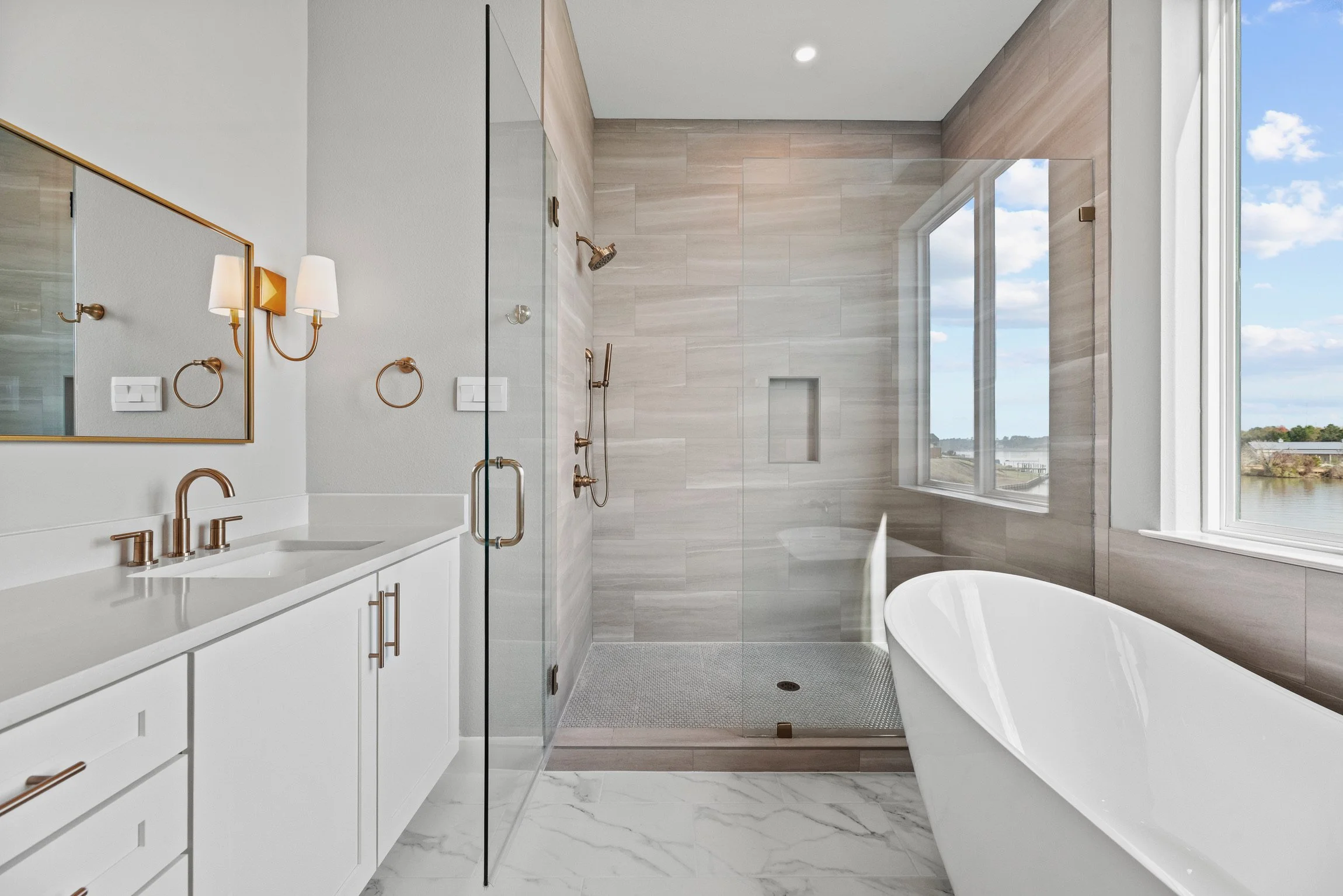 Modern bathroom with white vanity, gold fixtures, large window, freestanding bathtub, and glass-enclosed walk-in shower with beige tile walls.