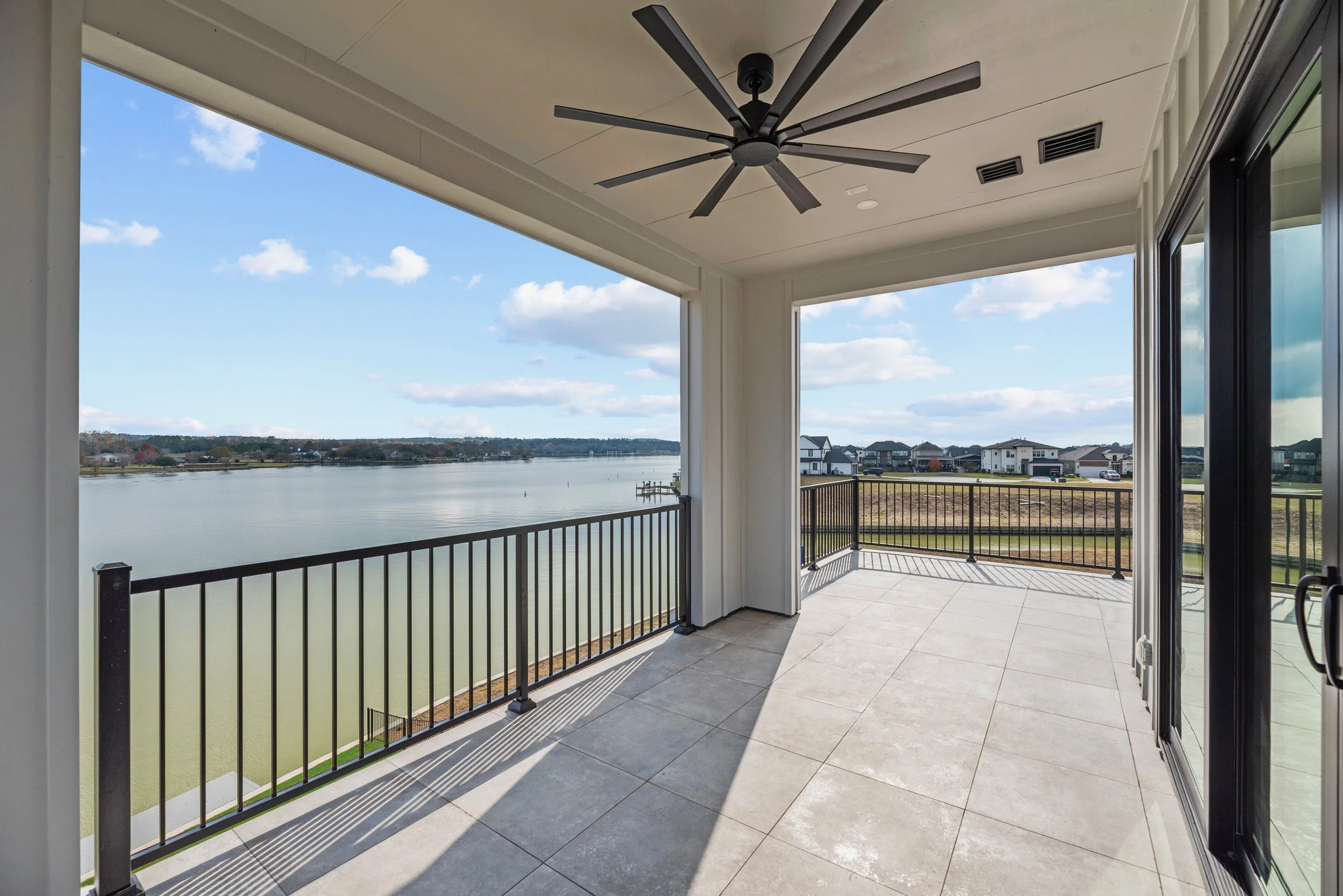 Empty covered balcony with black railing overlooking a lake and suburban houses under a partly cloudy sky.