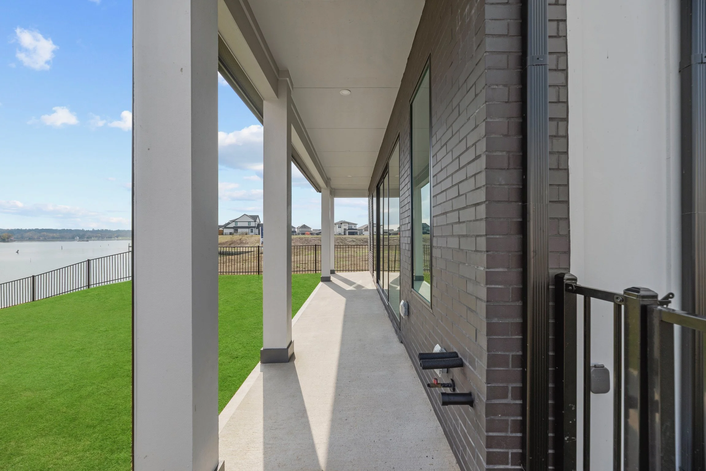 Side view of a modern house with a patio, glass sliding doors, brick and smooth wall exterior, overlooking a grassy yard and a body of water with houses in the distance under a partly cloudy sky.