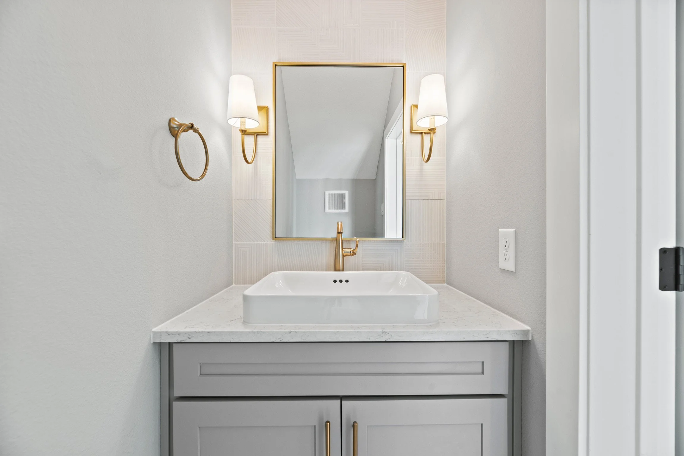 Modern bathroom vanity with white countertop, ceramic vessel sink, gold faucet, large mirror, wall-mounted gold light fixtures, beige tiled accent wall, and light gray cabinetry with gold handles.