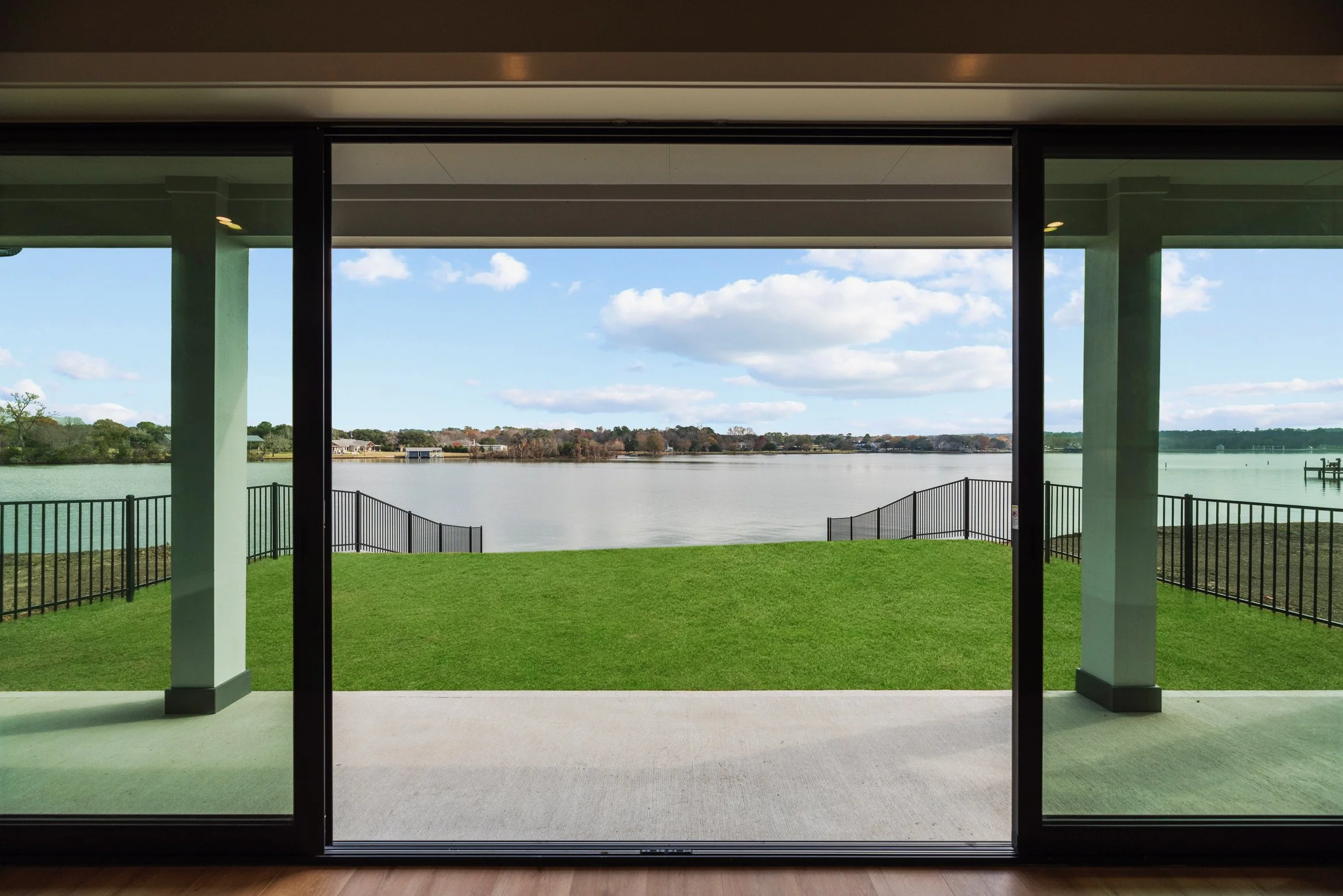 View of a lake with a grassy patio in the foreground, landscaped with black metal fencing, under a partly cloudy sky, seen from inside a room through large glass sliding doors.