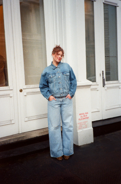 Young woman with curly hair wearing a denim jacket and wide-leg jeans standing outside near a white building with large windows and a door.